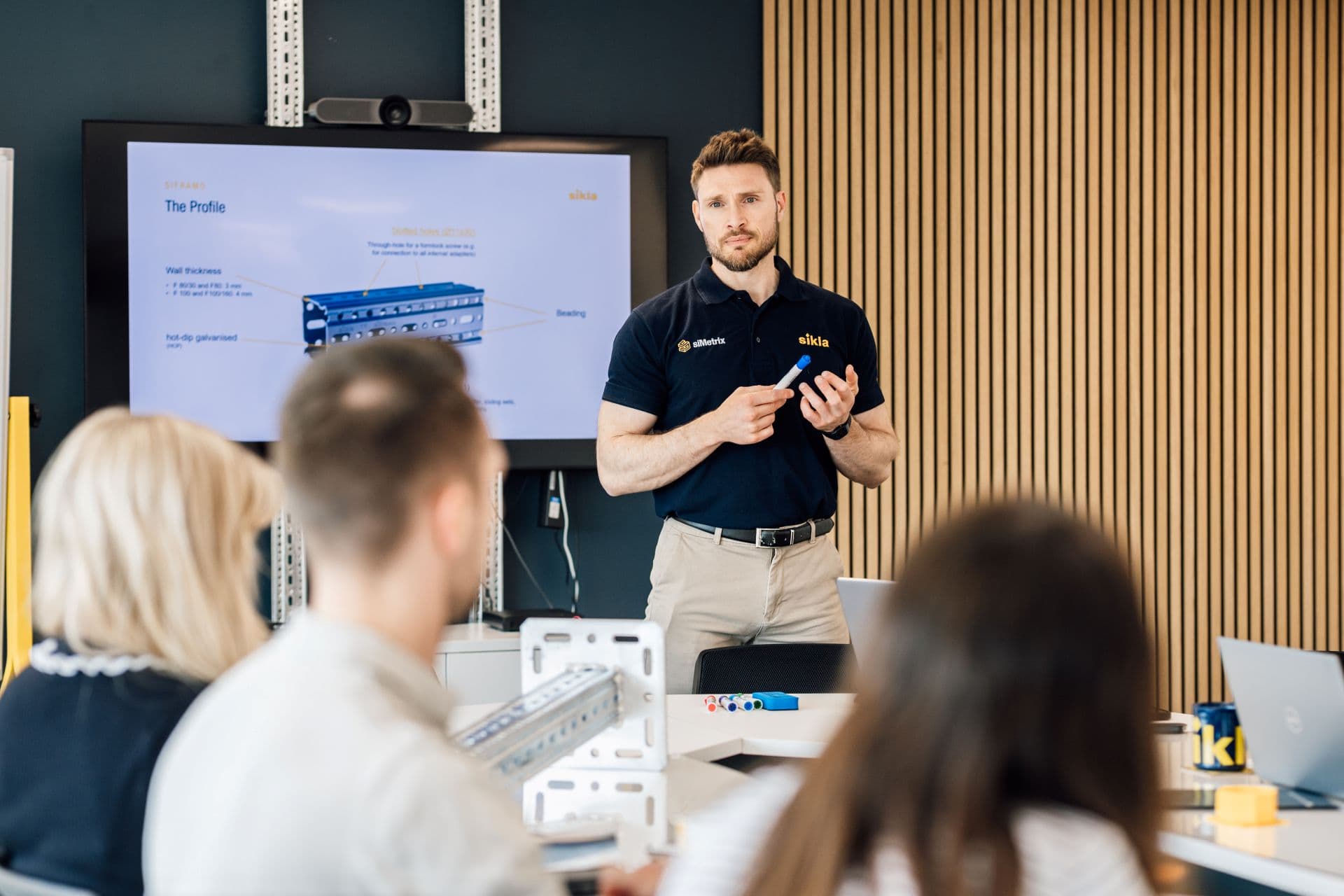 Man in navy polo holding a marker presenting a technical diagram to seated colleagues in a modern meeting room.